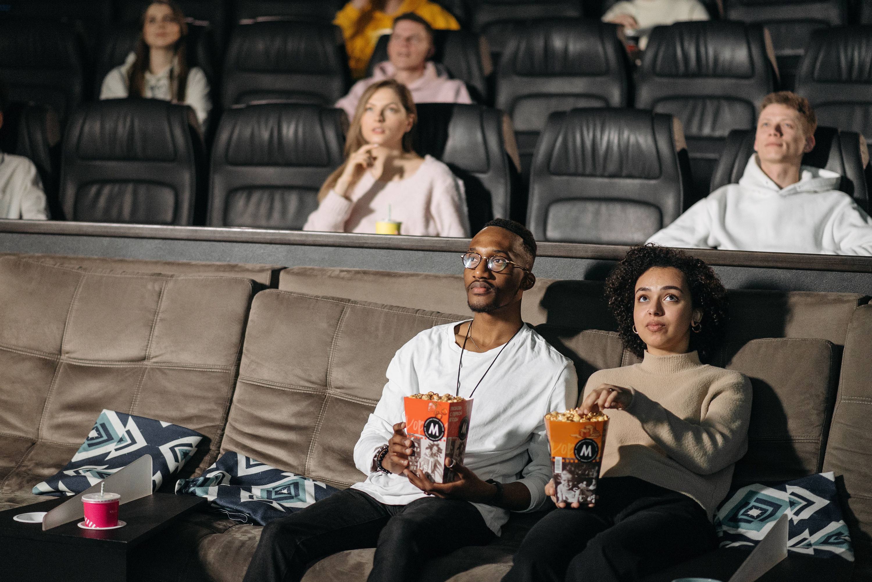 A young couple sitting in plush movie theater seats eating popcorn while watching a movie with other theatergoers.